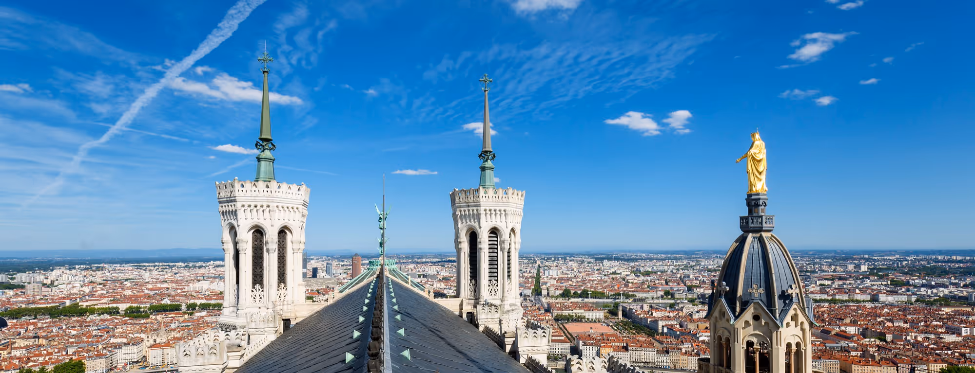 Vue panoramique de la Basilique Notre-Dame de Fourvière avec ses tours et la statue dorée dominant la ville de Lyon sous un ciel bleu.