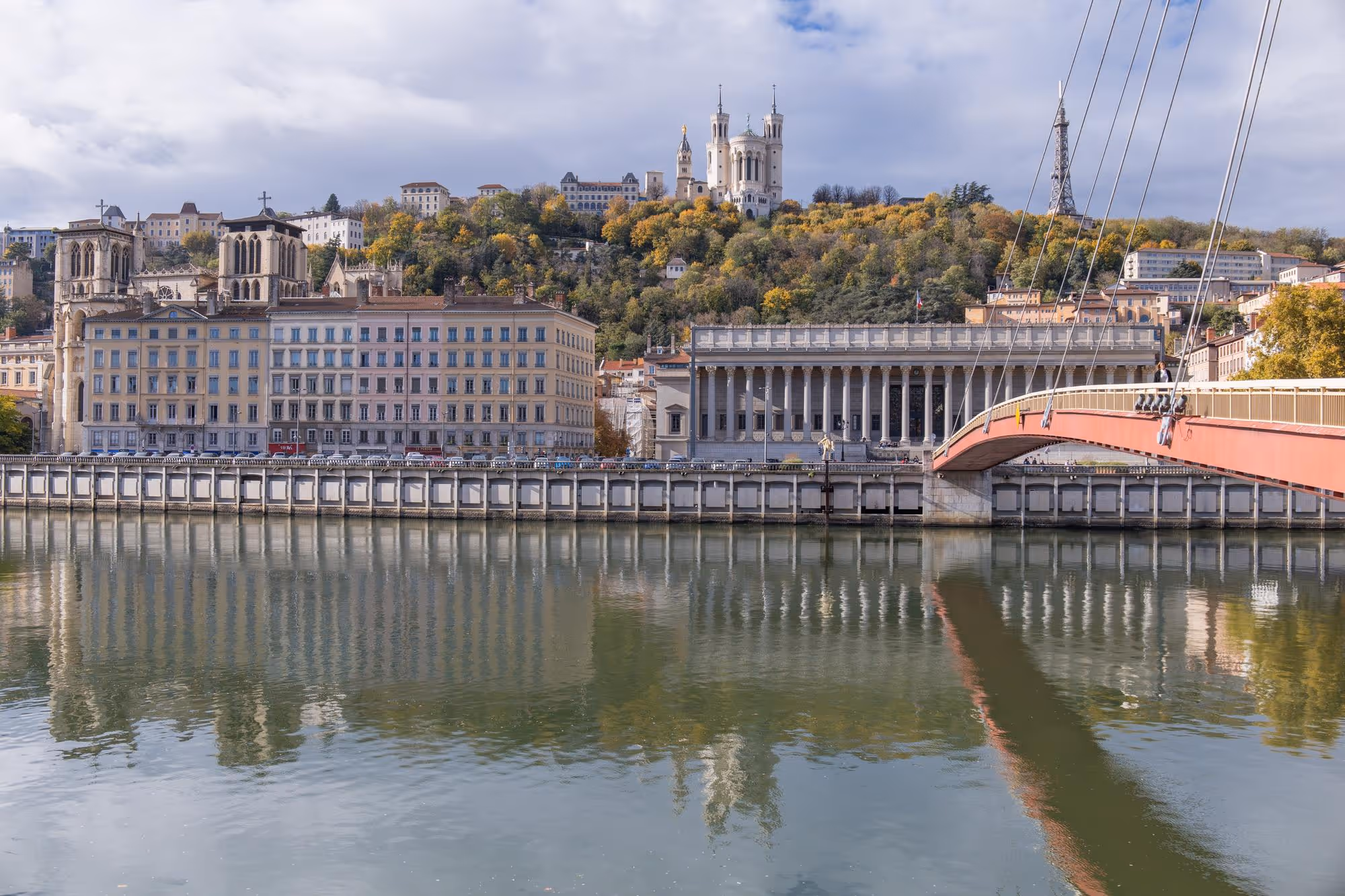 Vue panoramique du fleuve avec un pont suspendu rouge, des bâtiments historiques et la basilique de Fourvière sur une colline boisée à Lyon.