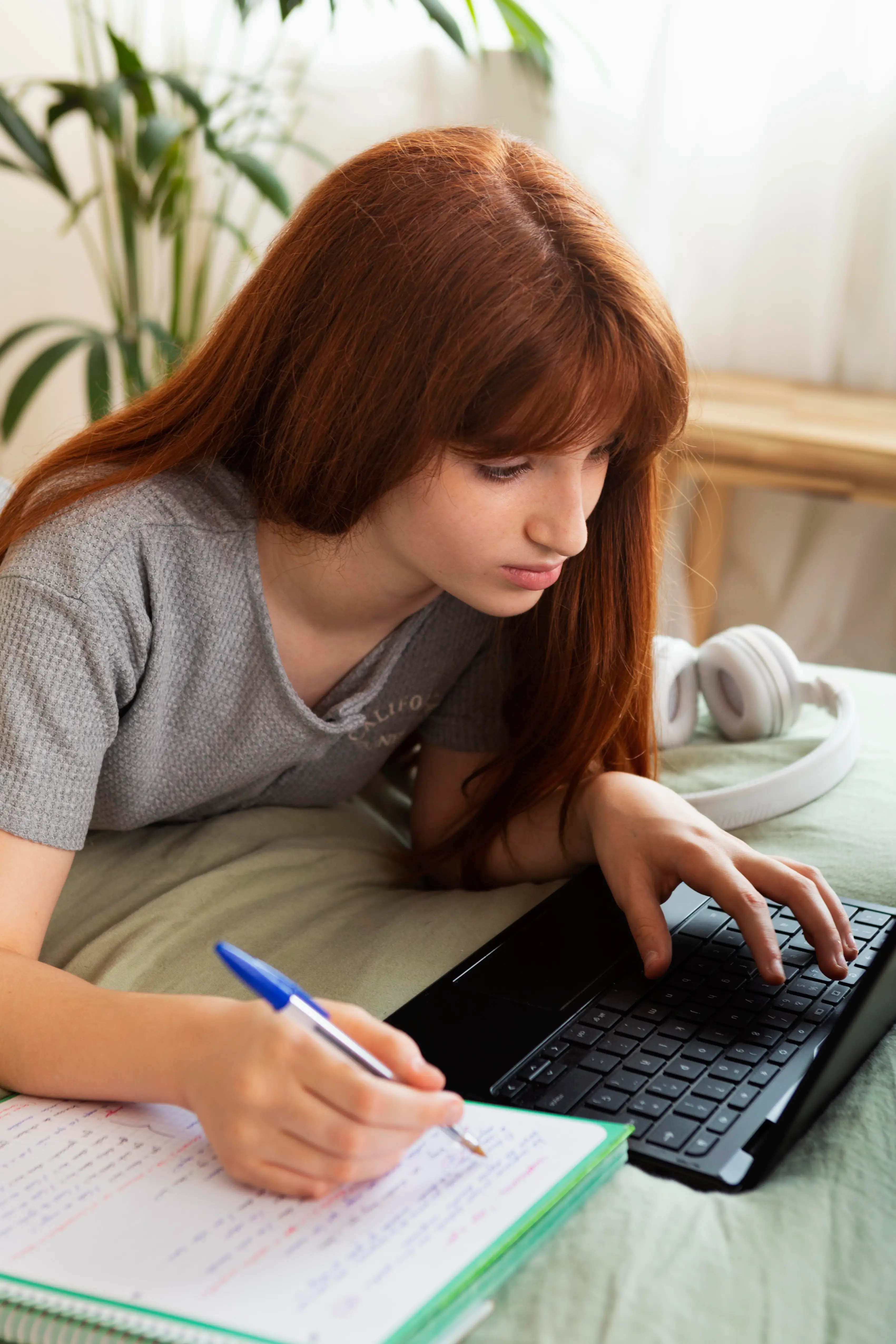 Dívka s načervenalými vlasy leží na posteli a píše do sešitu při používání klávesnice notebooku.