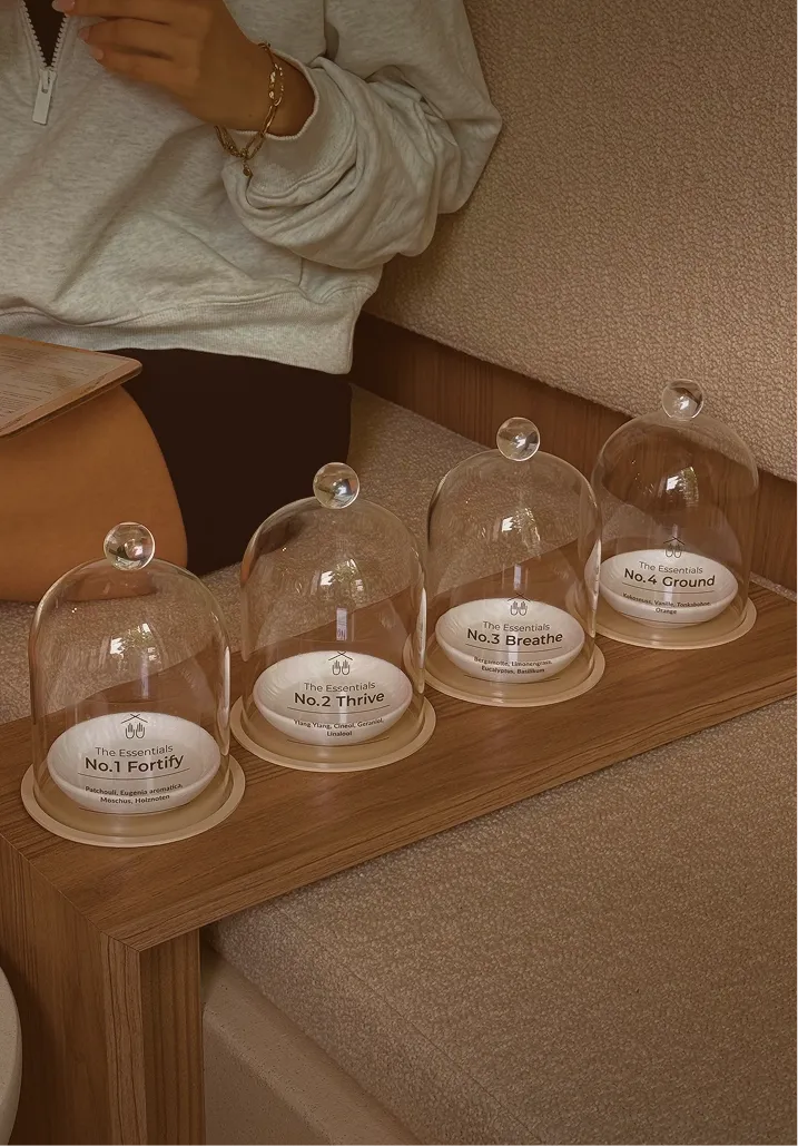 Vier Glasglocken auf einem Holztablett beschriftet mit den Essentials No.1 Fortify, No.2 Thrive, No.3 Breathe und No.4 Ground, jede mit unterschiedlichen Duftnoten.