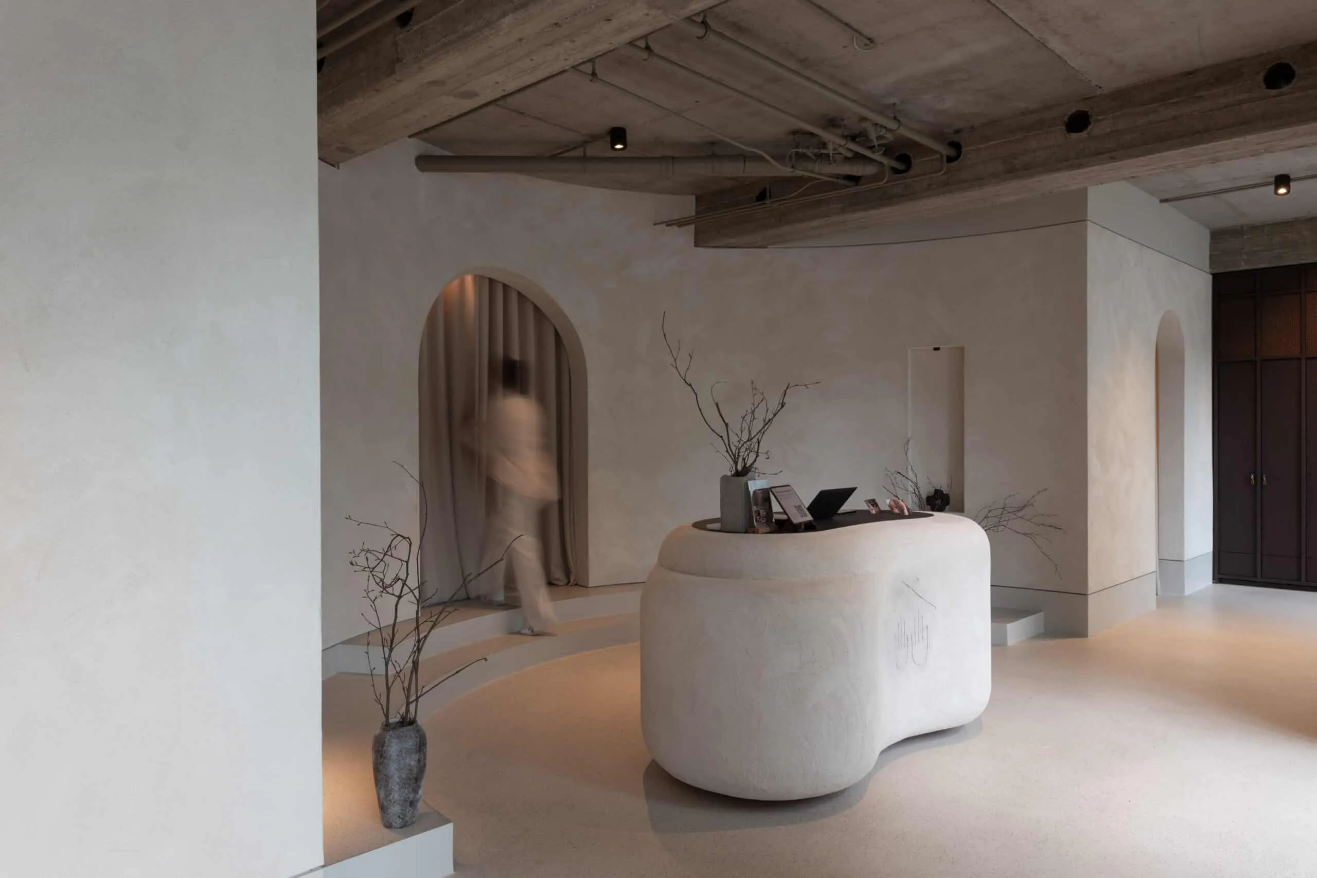 Moderner Empfangsbereich in einem Soulhouse Studio mit organisch geformtem weißen Tresen, kahlen Zweigen in Vasen und einer Person, die durch einen Vorhang geht.