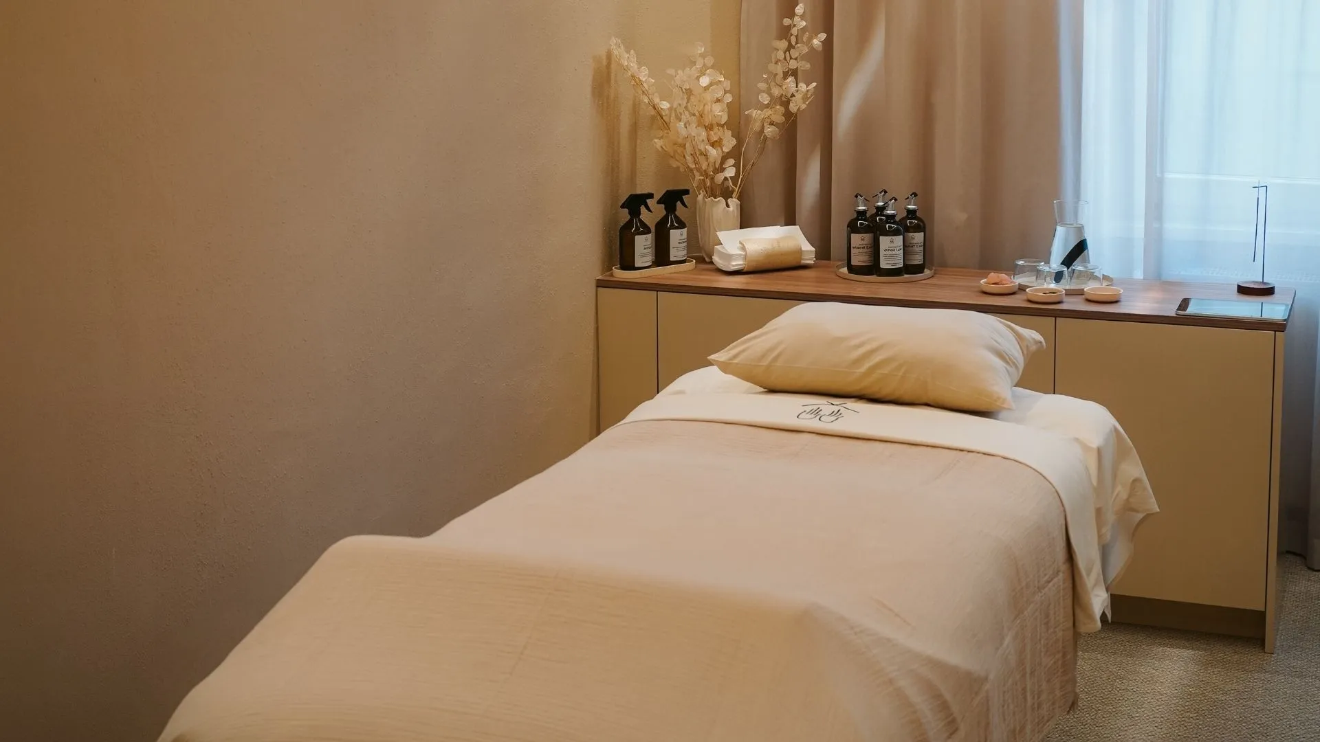 Soulhouse Studio Behandlungsraum mit massagetisch, beigem Leinenbezug und Kopfkissen vor einem Holzmöbel mit dekorativen Flaschen und getrockneten Blumen.