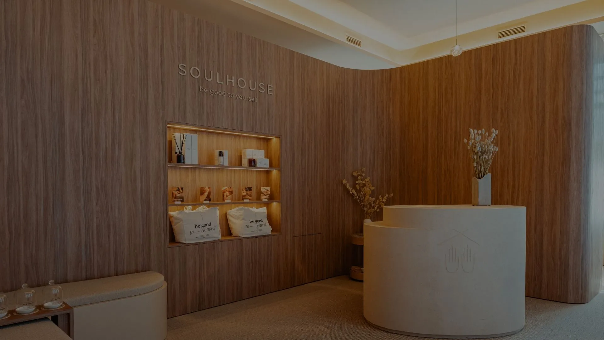 Empfangsbereich eines Soulhouse Studios mit einer runden weißen Rezeption, Holzwand mit beleuchtetem Regal und zwei Kissen mit der Aufschrift 'be good to yourself'.