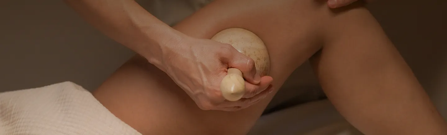 Hand hält ein hölzernes Massagegerät und führt eine Anti-Cellulite-Massage an einem Oberschenkel durch.