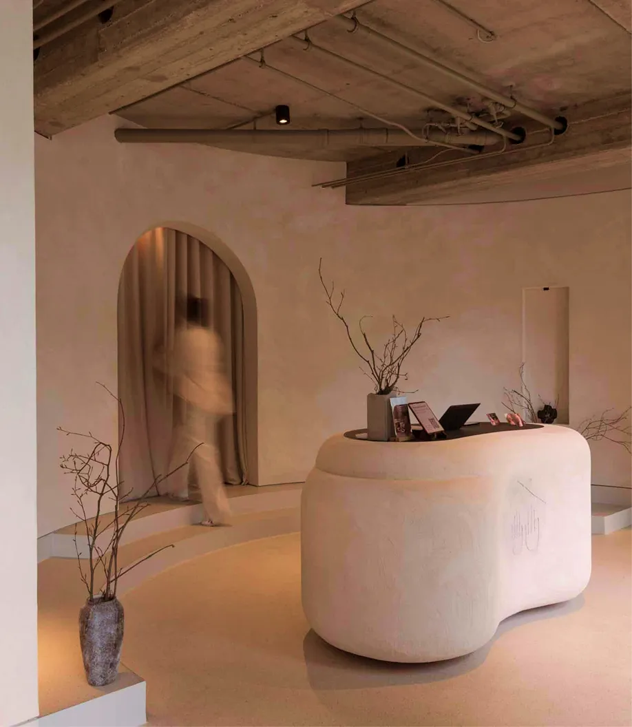 Minimalistischer Empfangsbereich eines Soulhouse Studios mit geschwungenem weißen Tresen, dekoriert mit Zweigen in Vasen, und einem verschwommenen Menschen vor einem bogenförmigen Vorhang.