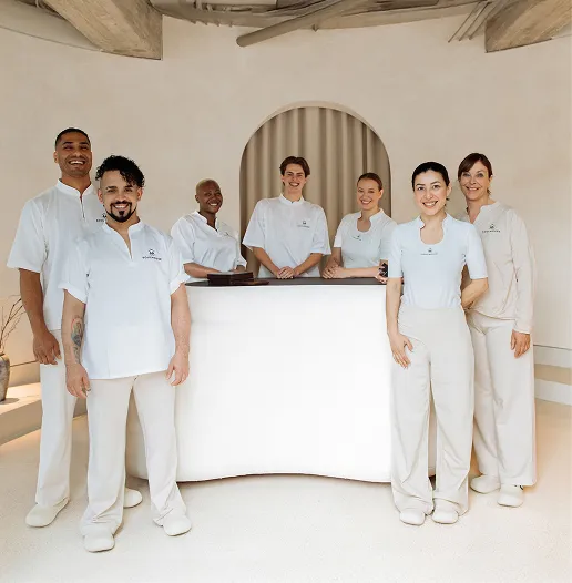 Teamfoto von sieben Soulhouse Massage Experten in weißen Klamotten in einem modernen, hellen Raum vor einem Empfangstresen.