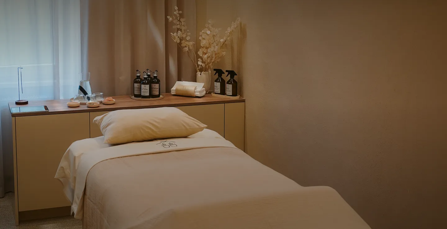 Ruhiger Behandlungsraum mit Liege, auf der ein Kissen liegt, ein Tisch mit Aromatherapie-Ölen, Handtüchern und getrockneten Blumen in einer Vase.