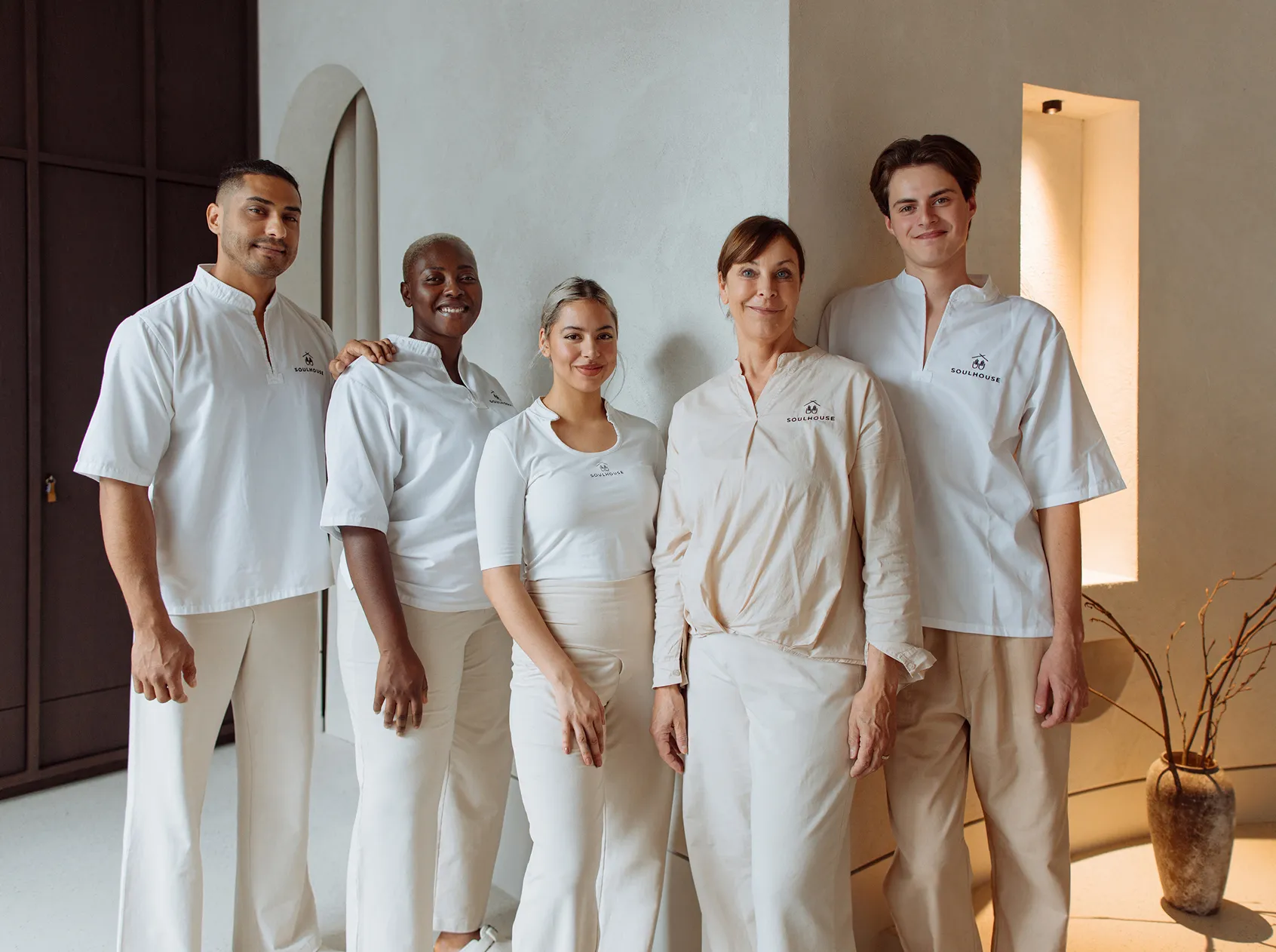 Gruppenbild von 5 Massage Experten in weißen und beigen Uniformen mit dem Soulhouse-Logo posieren in einem hellen Raum mit einer Vase im Hintergrund.
