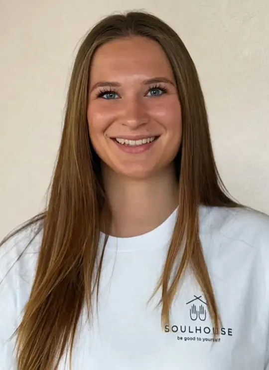 Soulhouse Massage Expertin mit langem braunem Haar lächelt, trägt ein weißes T-Shirt mit dem Logo und Schriftzug Soulhouse.