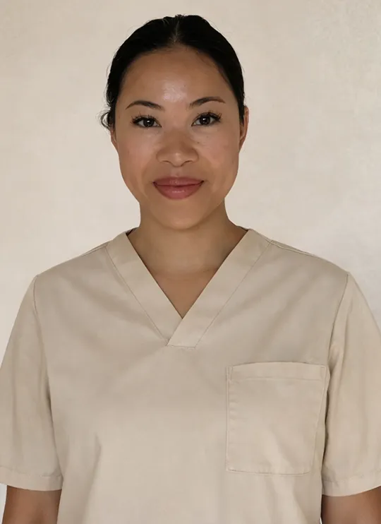 Soulhouse Massage Expertin mit zurückgebundenem Haar, die beige medizinische Arbeitskleidung trägt und freundlich lächelt.