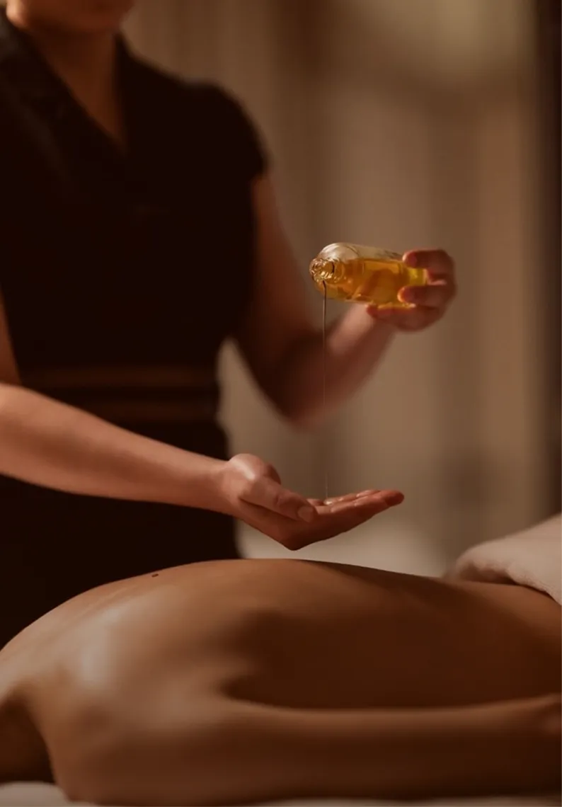 Eine Person gießt Massageöl auf ihre Hand über dem Rücken einer liegenden Person in einem warm beleuchteten Raum.