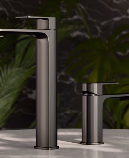 Two modern matte black faucets of different heights installed on a white marble countertop with dark green plants and a dark marble wall in the background.