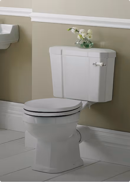 White vintage-style toilet with a closed lid and a small vase with white flowers on the tank in a beige bathroom.