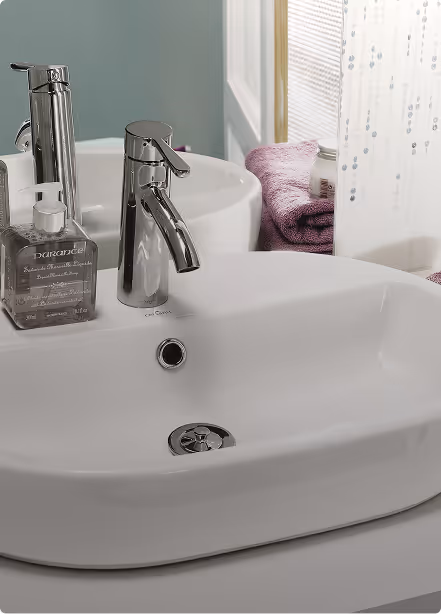 Modern white bathroom sink with chrome faucet and clear soap dispenser on the left side, purple towel and shower curtain in the background.