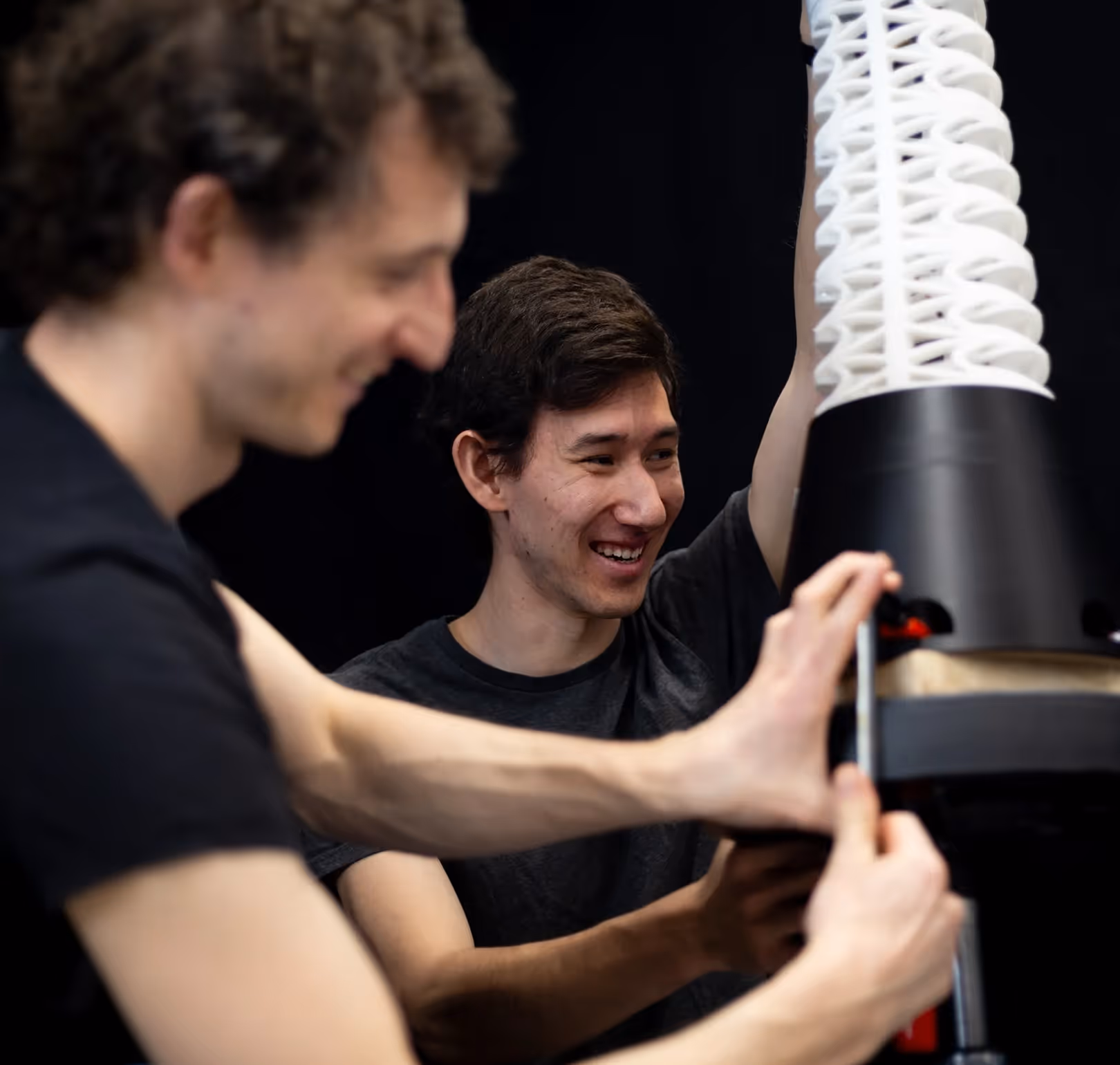 Two men smiling and working together on assembling a tall, white and black mechanical or robotic device.