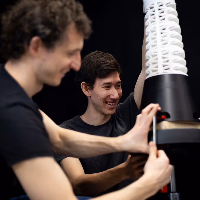 Two young men collaborating on assembling a structure with a black base and a white, lattice-like vertical component.