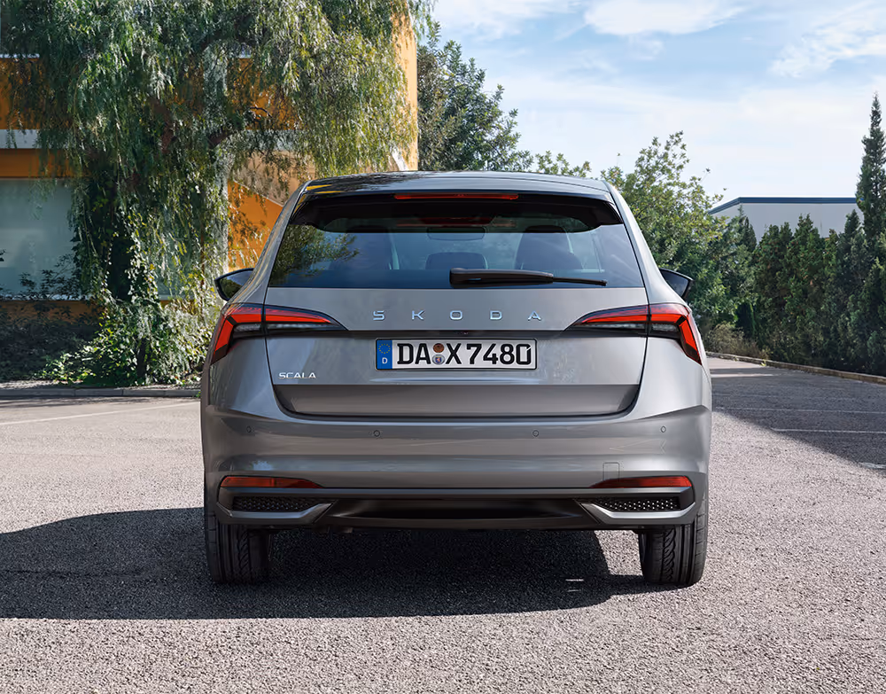 Heckansicht eines Škoda Scala Balance in der Lackierung Graphit-Grau Metallic auf einem Parkplatz vor einer grünen Baumkulisse. Zentral auf der Heckklappe ist der Škoda Schriftzug platziert, eingerahmt von den charakteristischen LED-Heckleuchten. Der untere Bereich des Stoßfängers rundet die funktionale Ästhetik des Kompaktmodells ab.