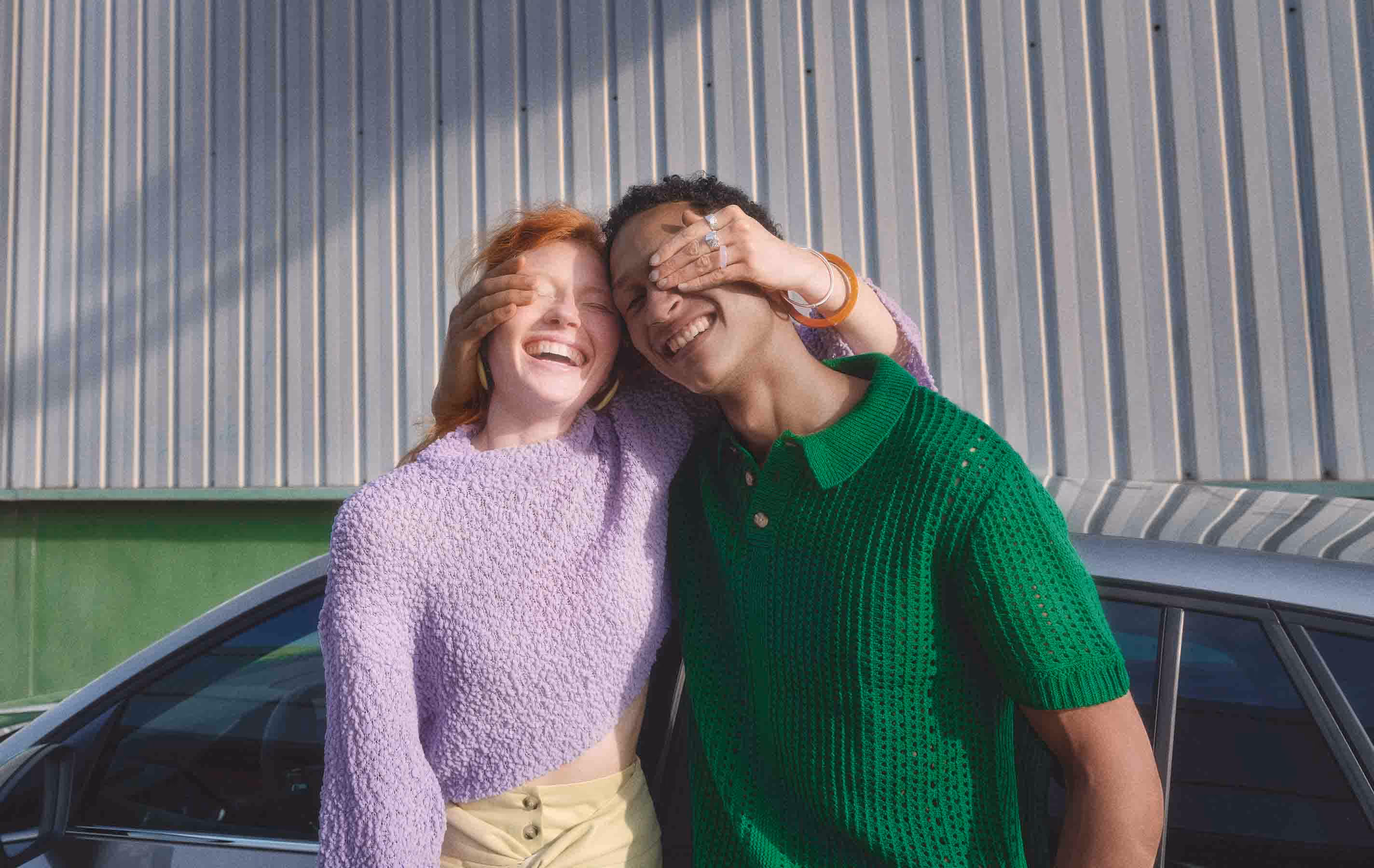 Lifestyle-Bild zweier lachender Personen, die sich neben einem Škoda Scala Balance spielerisch gegenseitig die Augen zuhalten. Von dem Fahrzeug ist lediglich ein Ausschnitt der Dachpartie und der Seitenfenster zu erkennen. Die Szene spielt sich vor einer hellen Industriewand ab.