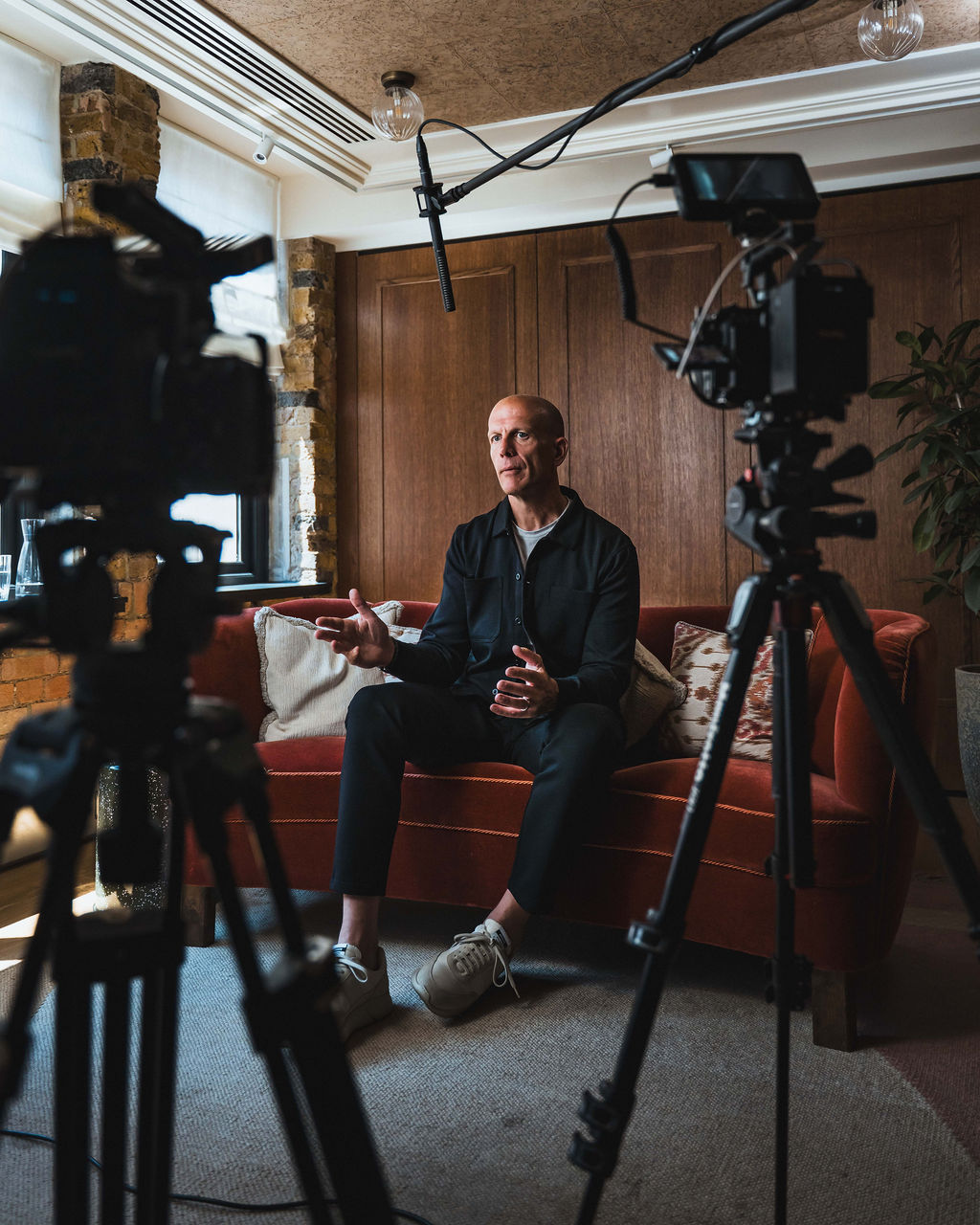 Man sitting on a red sofa speaking during a video interview with cameras and a boom microphone set up in front of him.