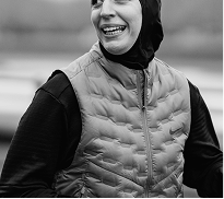 Smiling person wearing a hijab and quilted vest outdoors.