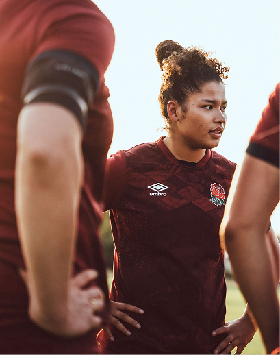 Woman rugby player in maroon jersey with hands on hips standing among teammates during practice or game.