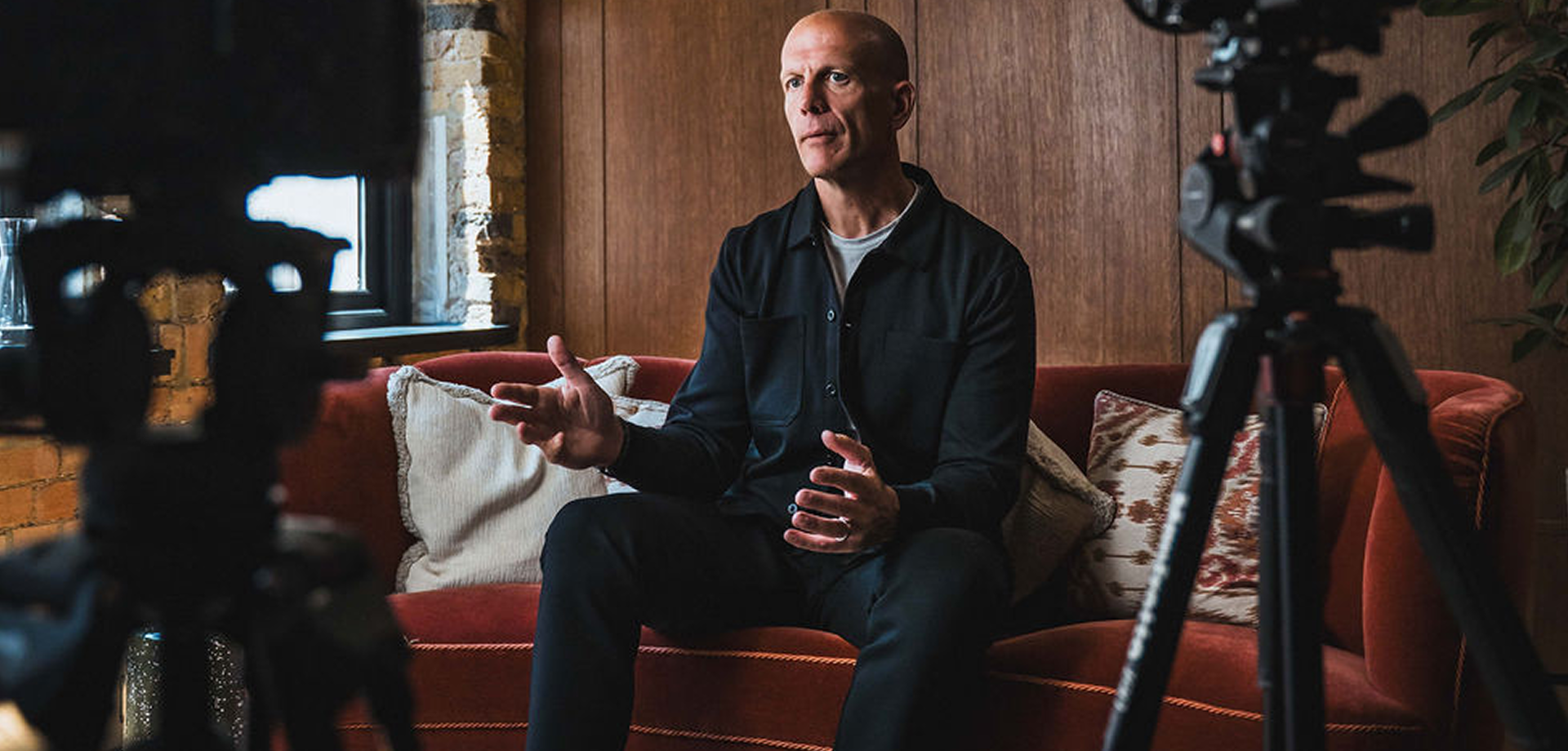 A bald man in a black shirt sitting on a red couch, gesturing with his hands during a video recording.