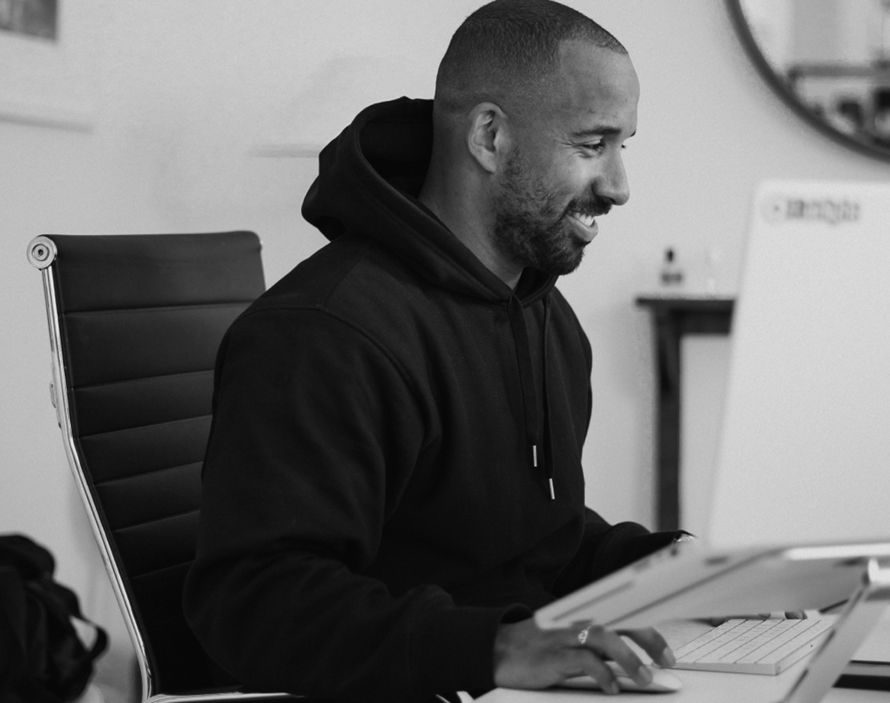 Smiling man in a black hoodie working on a laptop at a desk.