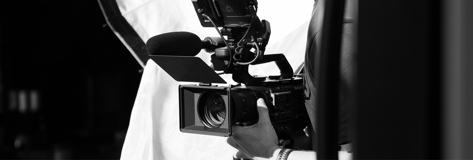 Bald man sitting and looking thoughtful during a video recording with cameras and lighting equipment in a room with wooden paneling and exposed brick.