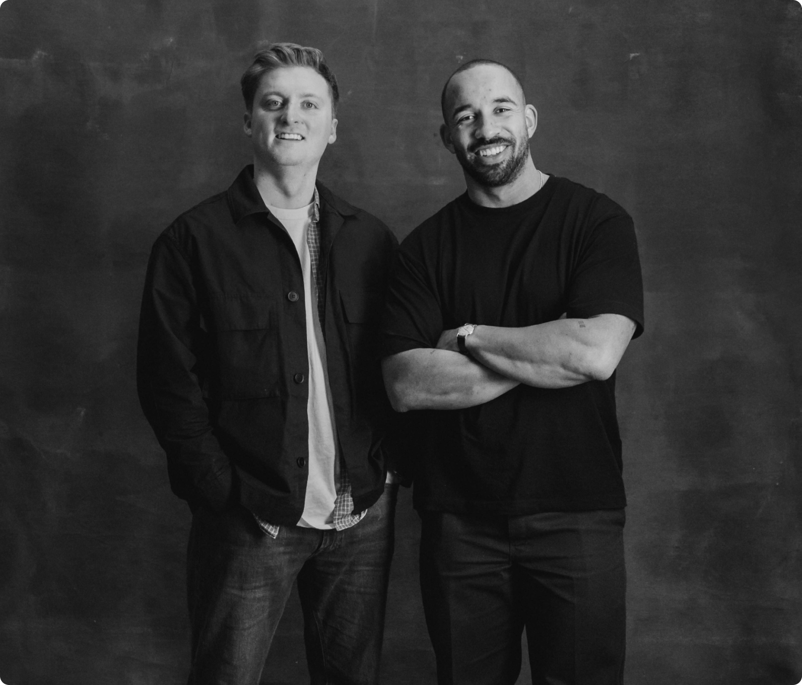 Two men standing side by side against a plain dark background, both smiling; one man has his hands in his pockets wearing a jacket, the other crosses his arms wearing a t-shirt.
