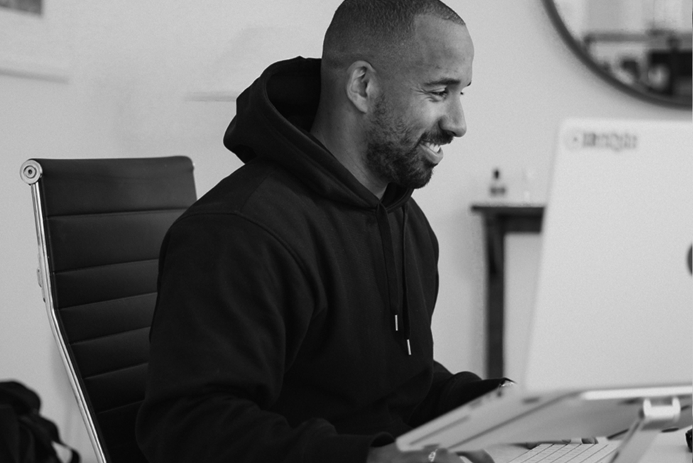 Smiling man in a black hoodie sitting on a chair working on a laptop.