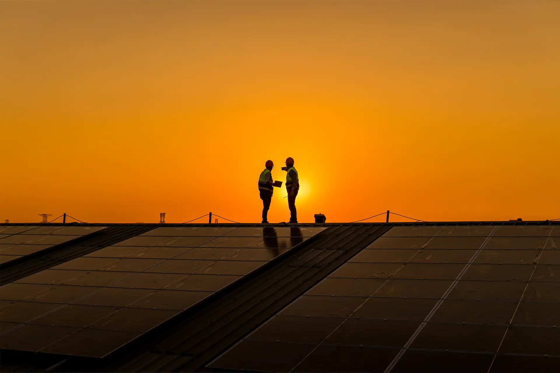 Deux techniciens en tenue de sécurité discutant sur un toit équipé de panneaux solaires au coucher du soleil.