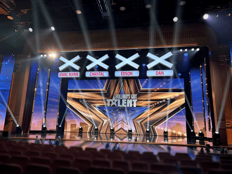 Podium van Holland's Got Talent met vier grote X-symbolen en namen van juryleden Marc-Marie, Chantal, Edson en Dan boven een verlicht toneel.