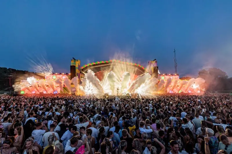 Groot publiek op een buitenmuziekfestival bij schemering met een verlicht podium en rook- en lichteffecten.