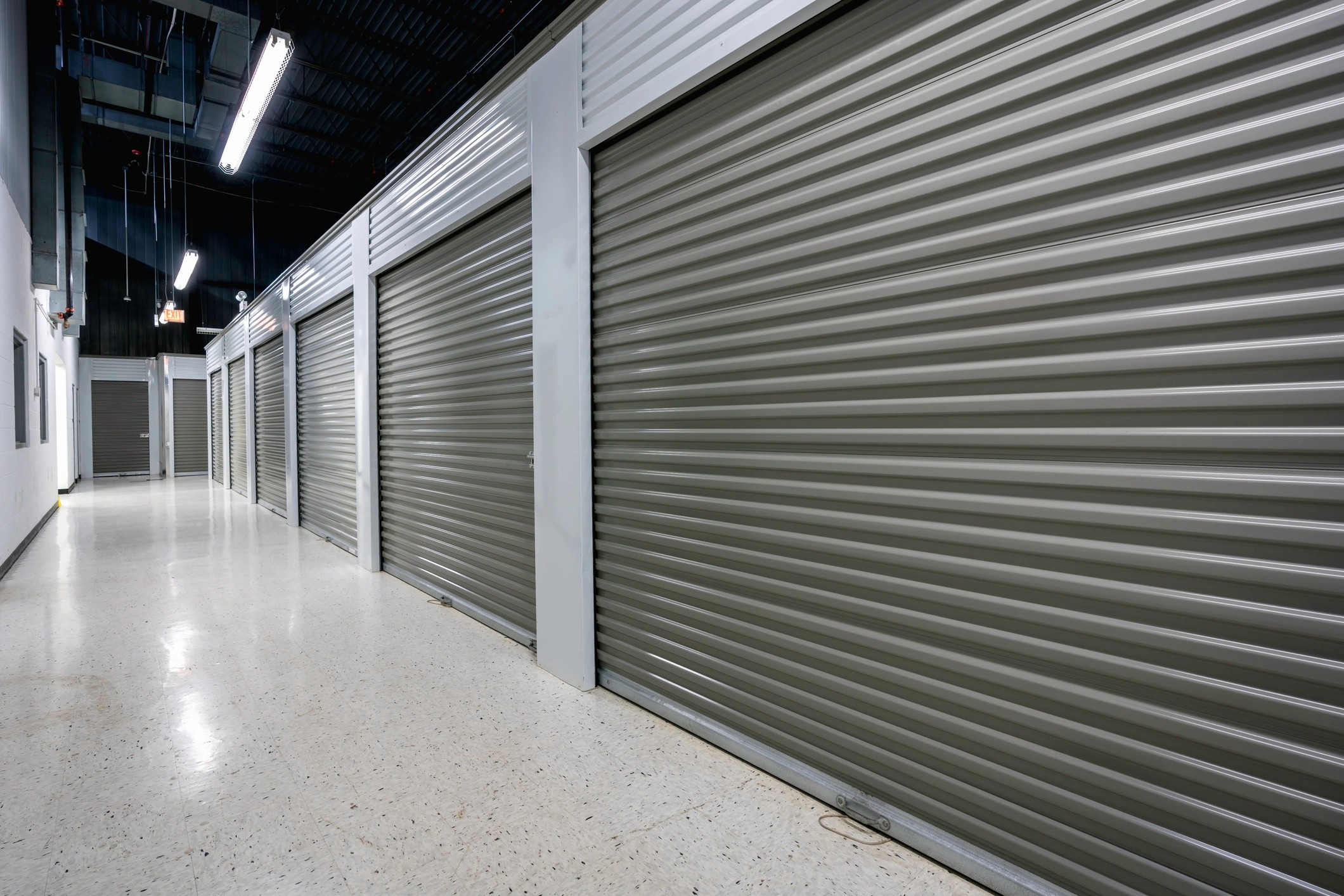 Interior hallway of a storage facility with multiple closed gray roll-up metal doors and white walls.