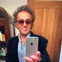 Man with curly hair and sunglasses smiling while taking a selfie with a smartphone in front of a wooden door.