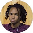 Portrait of a young person with short dreadlocks wearing a dark purple hoodie against a wooden background.