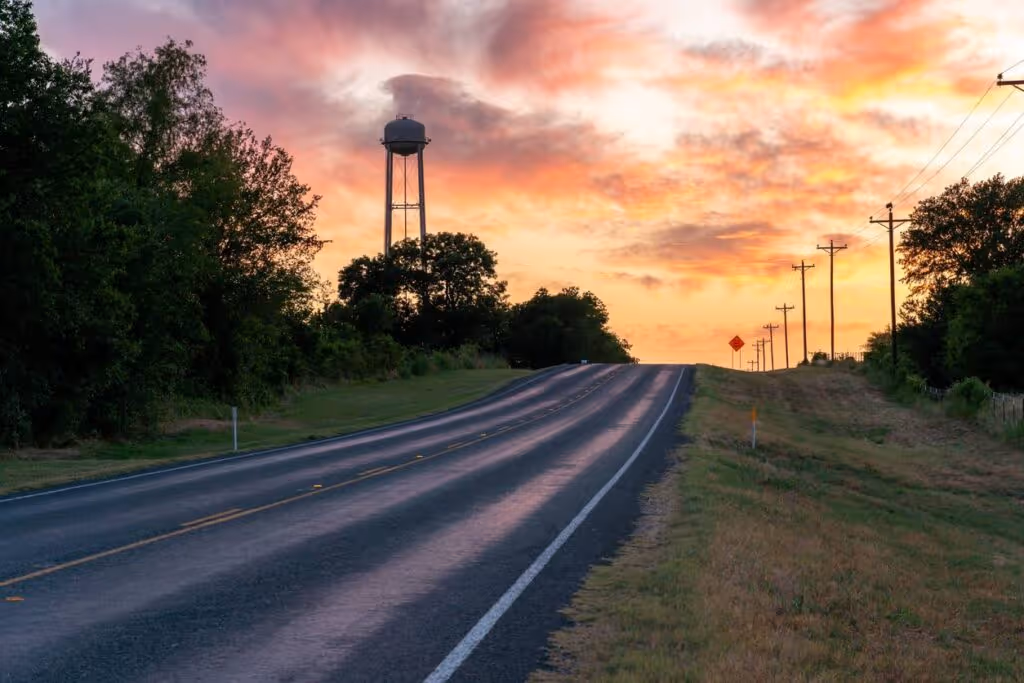 A road leading into the sunset surrounded by trees with a water tower’s silhouette.