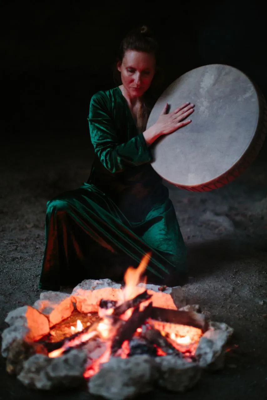 Woman in green dress playing a drum while kneeling near a campfire at night.