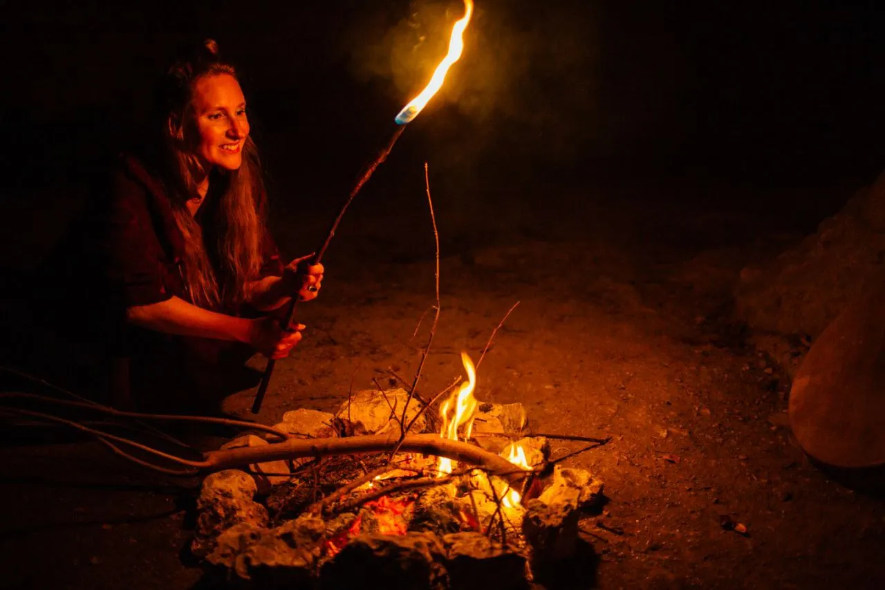 Smiling woman holding a torch near a campfire at night outdoors.