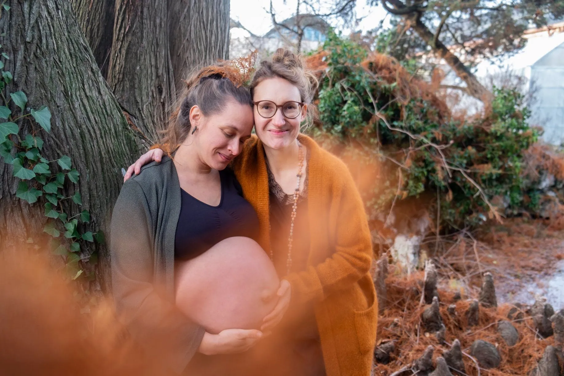 Two women outdoors, one pregnant with a bare belly, embracing by a large tree with autumn leaves.