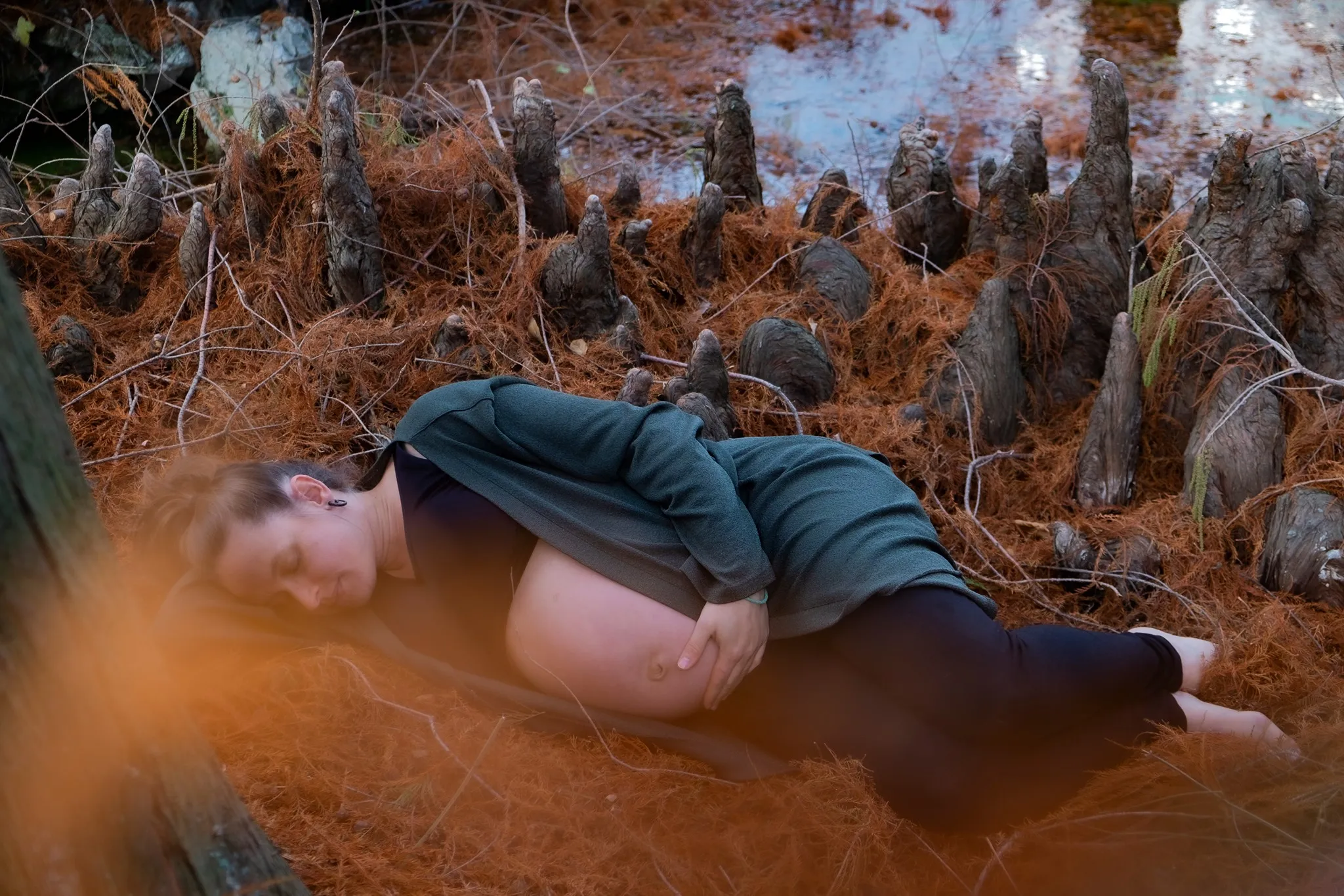 Pregnant woman lying on orange pine needles near cypress knees in a forest.