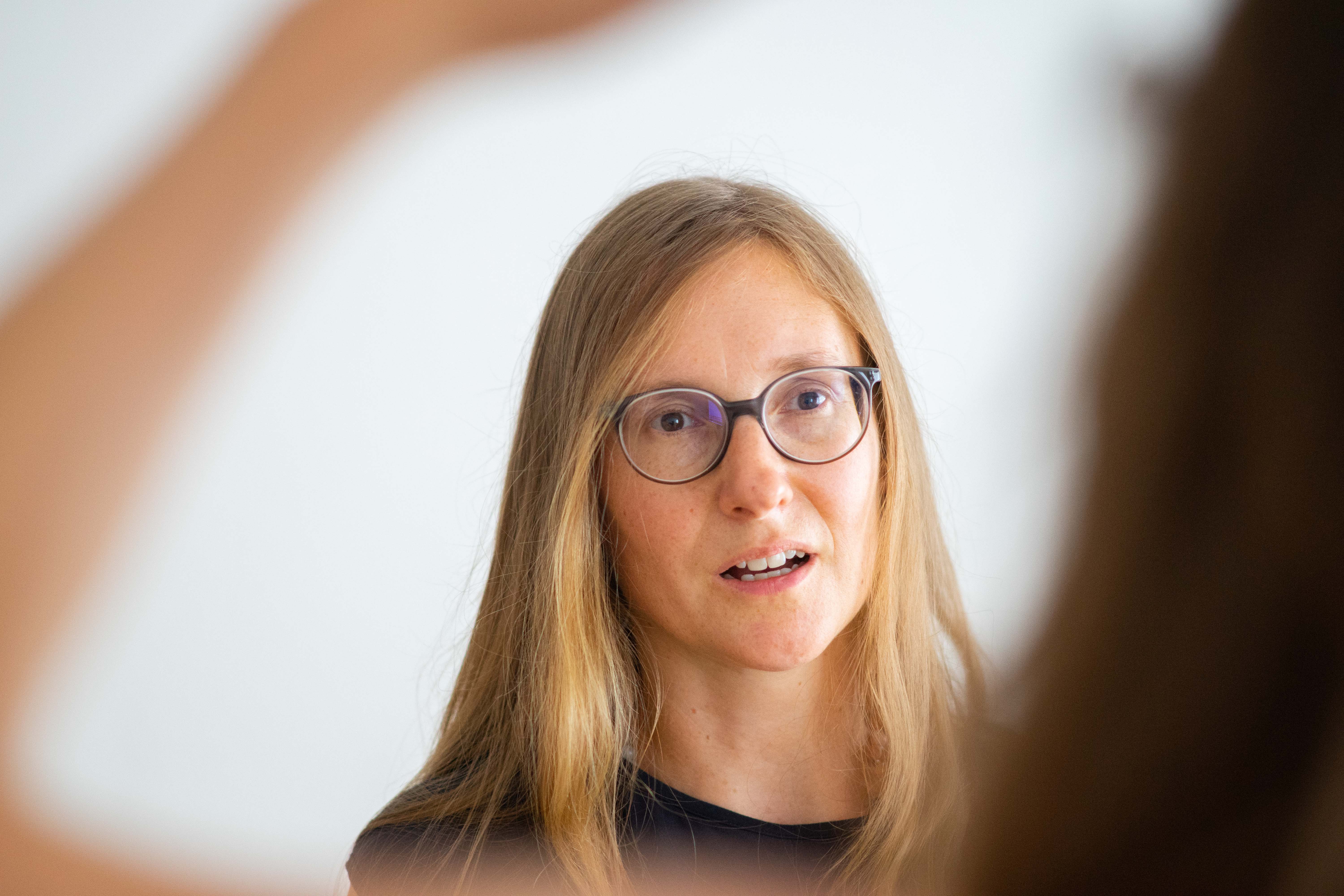 Woman with long blonde hair and glasses speaking, with blurred arms framing her face.