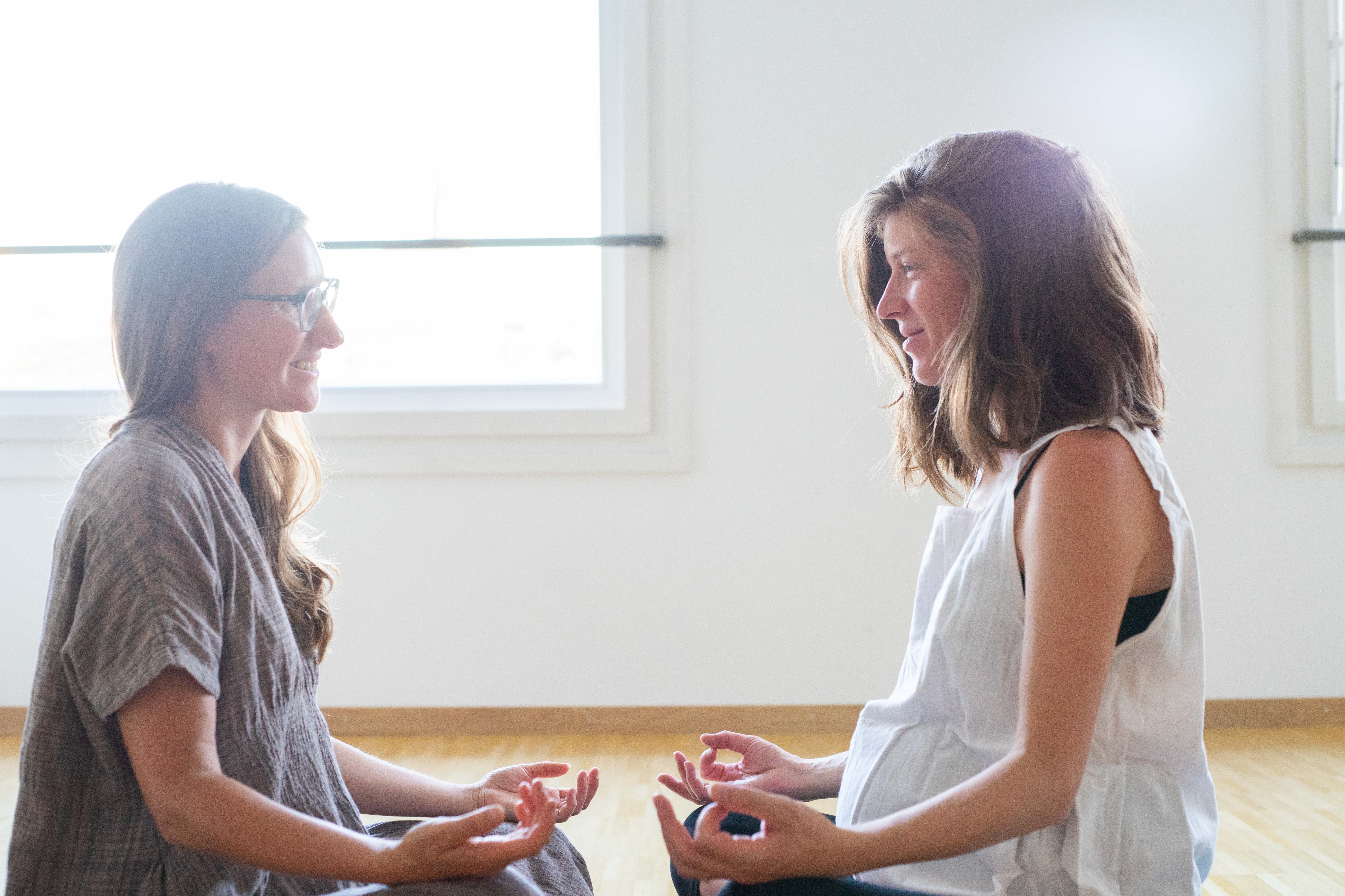 Two women sitting cross-legged on wooden floor facing each other with hands in meditation pose.