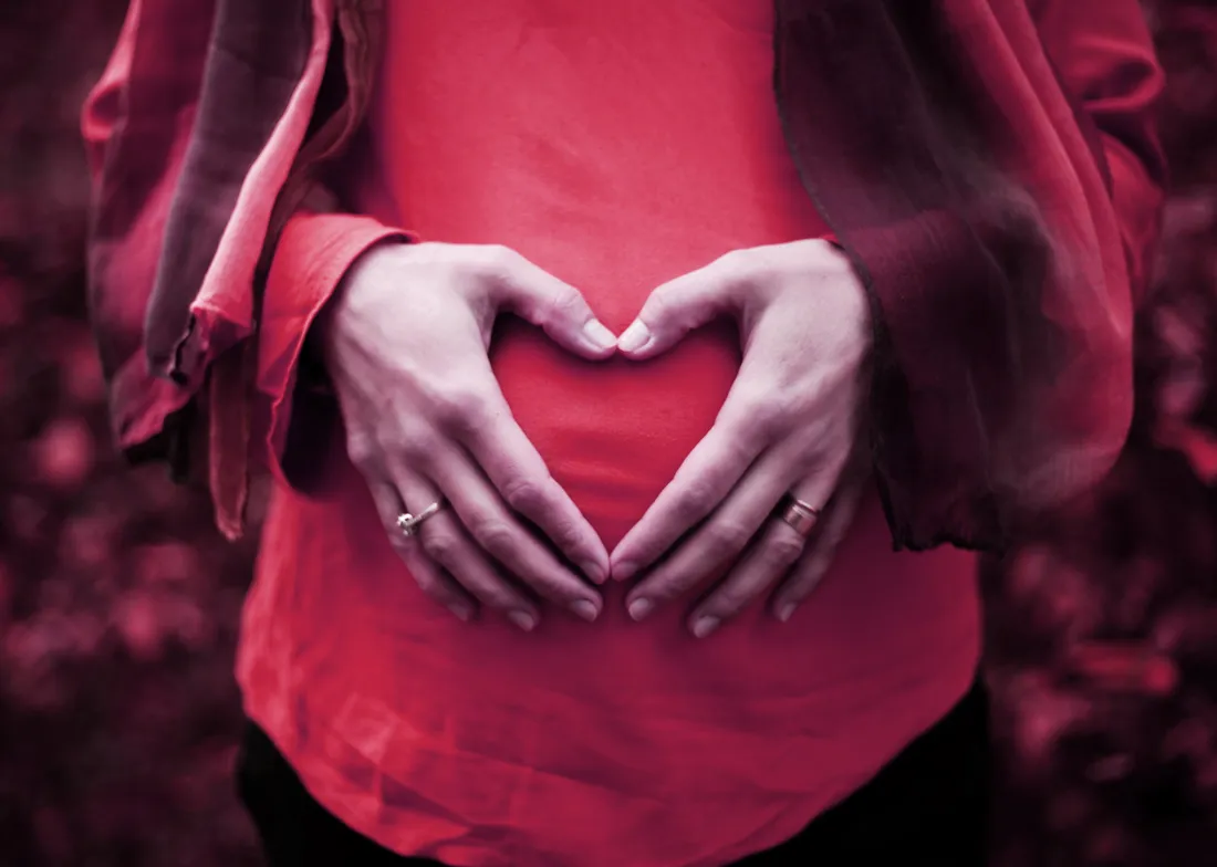 Person in red shirt making a heart shape with hands over their pregnant belly.