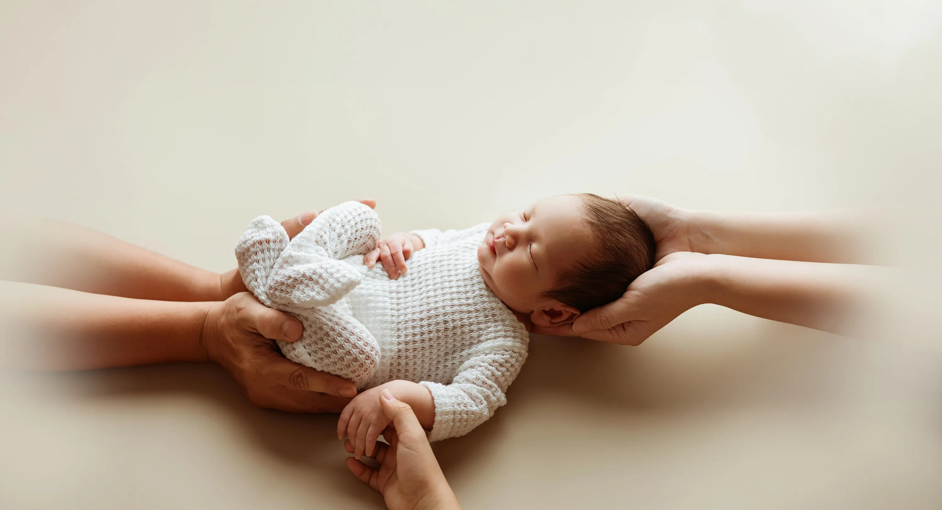 Newborn baby in white knit outfit held and supported by two adult pairs of hands on beige background.