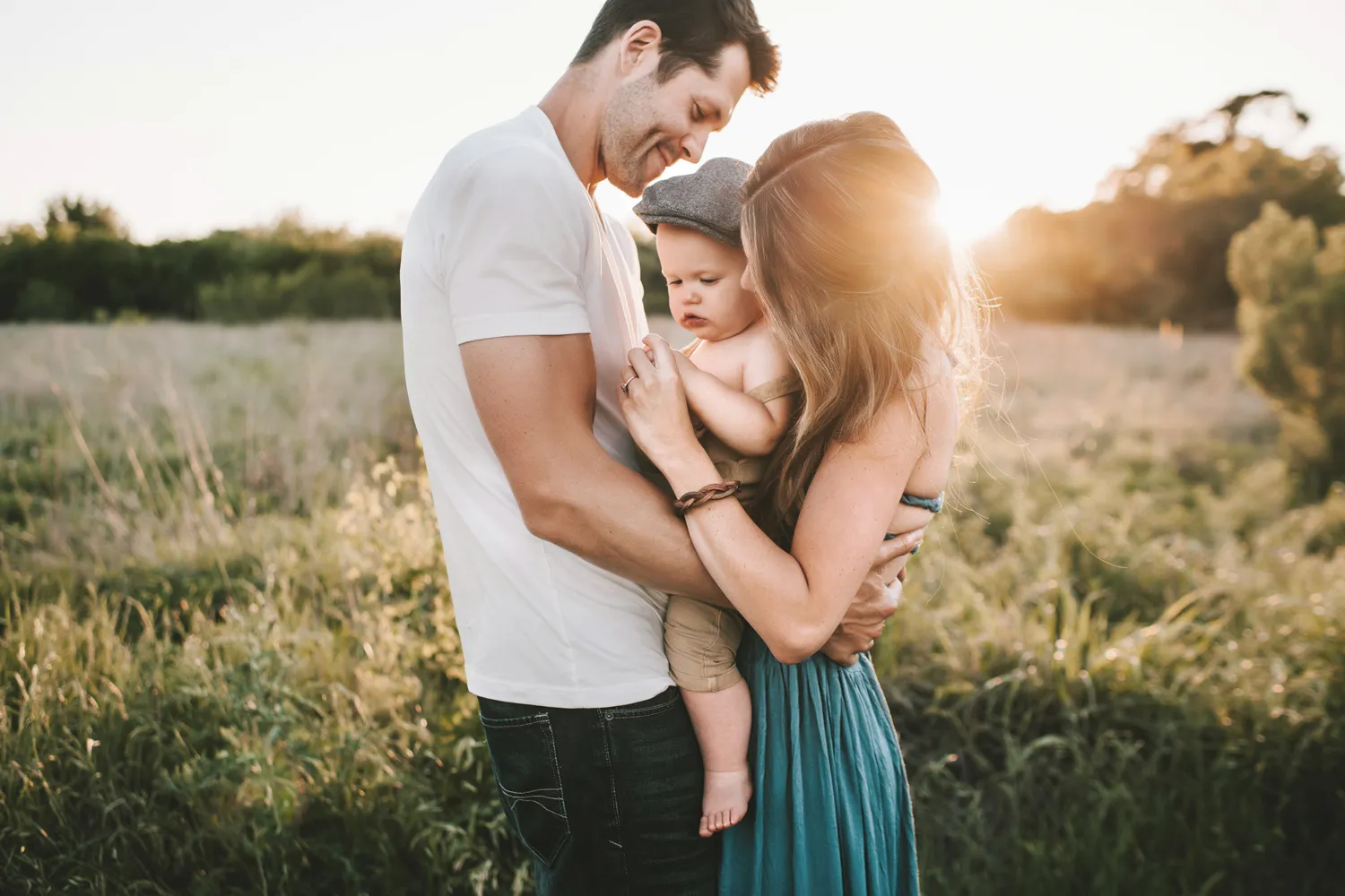 Man and woman holding a baby outdoors in a sunny grassy field during sunset.