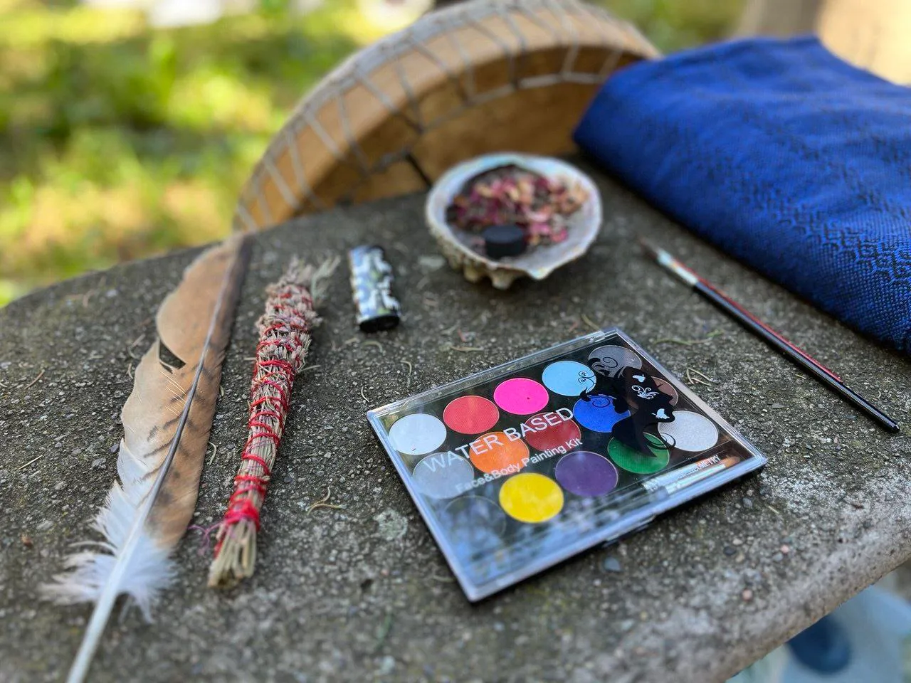 Face and body paint kit next to paintbrush, feather, smudge stick, shell with herbs, and blue cloth on stone table.