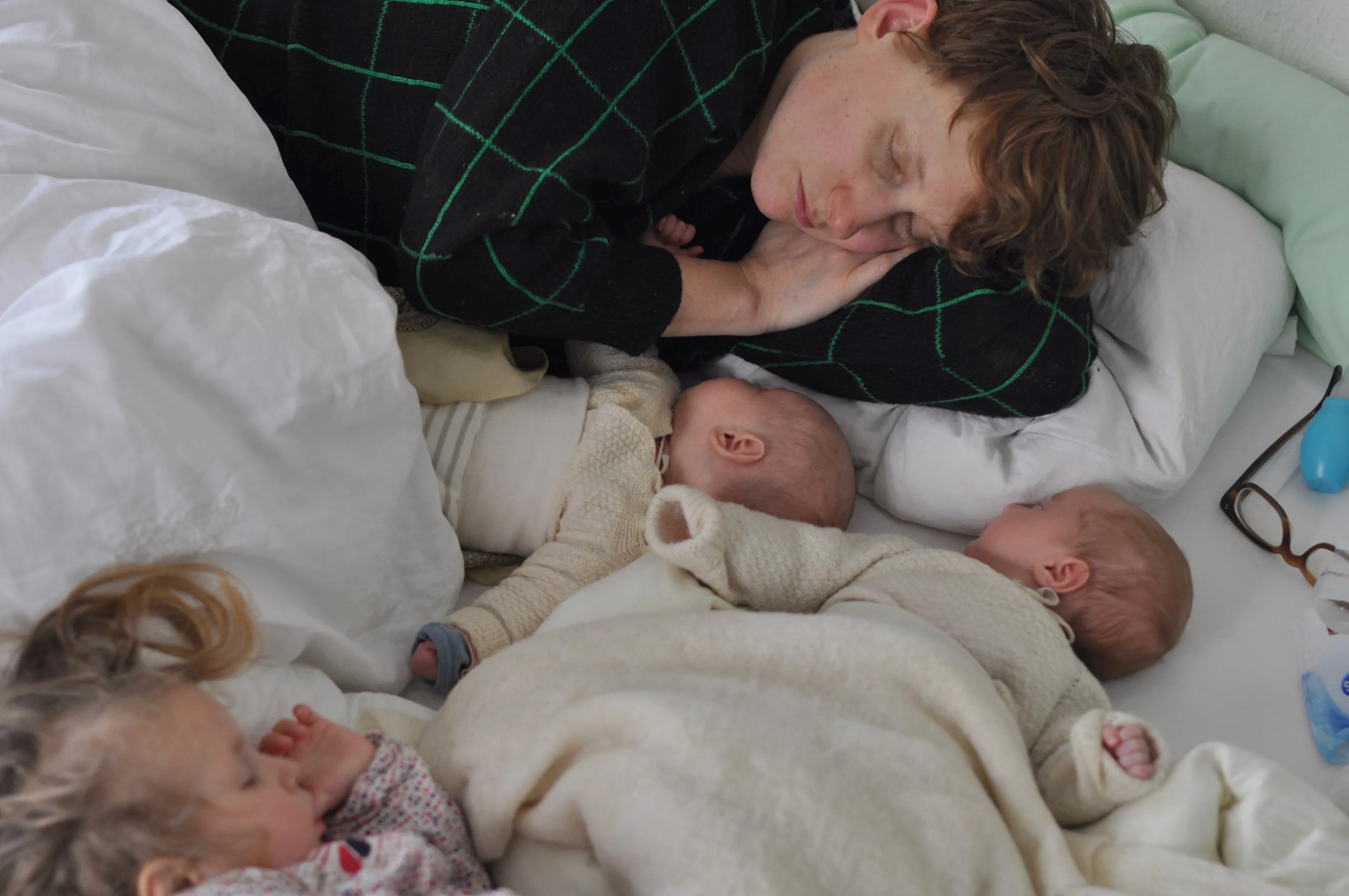 Sleeping adult with two babies and a toddler resting together on a bed with white bedding.