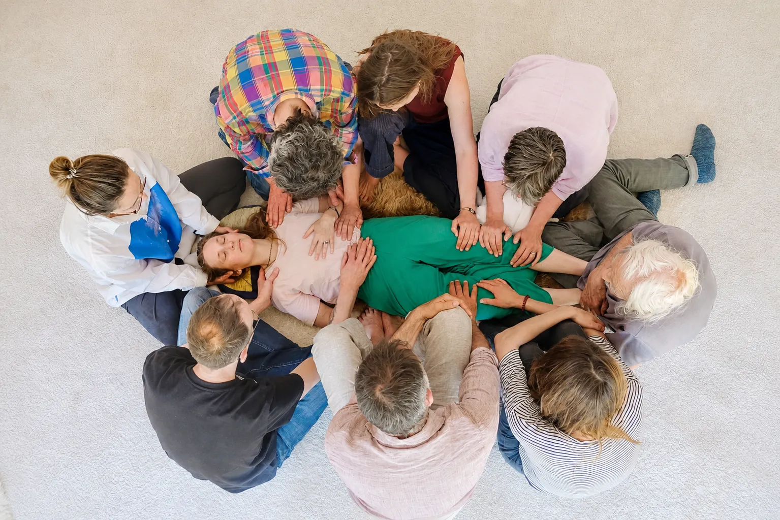 Group of eight people sitting in a circle gently touching a woman lying relaxed on the floor.