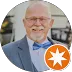 Smiling older man with glasses and beard, wearing a blue suit and orange tie.